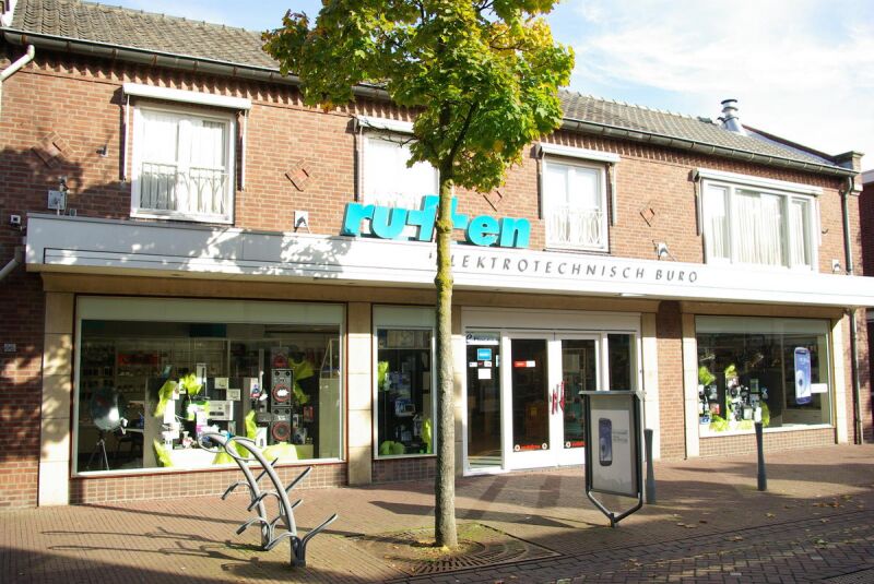 Vooraanzicht van de winkel van een elektrotechnisch bureau met grote etalages en een boom voor de ingang.