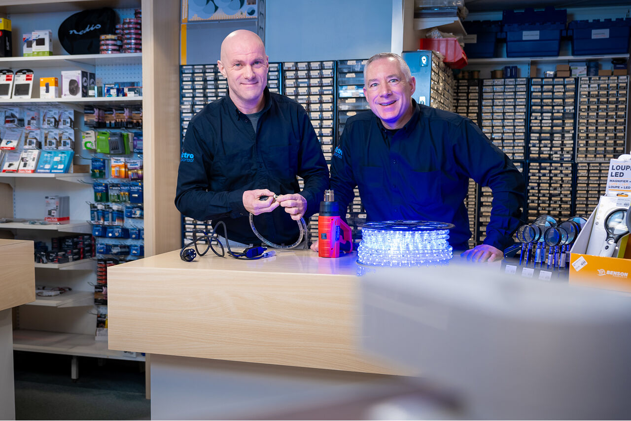 Twee medewerkers van Elektroportaal achter de toonbank met elektrische producten, waaronder een LED-lichtslang en gereedschap.
